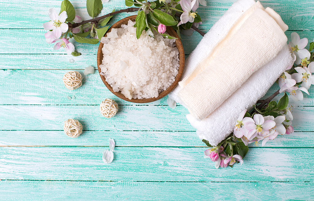 Spa or wellness setting. Sea salt in bowl, towels and apple blossom on turquoise wooden background. Selective focus is on salt.