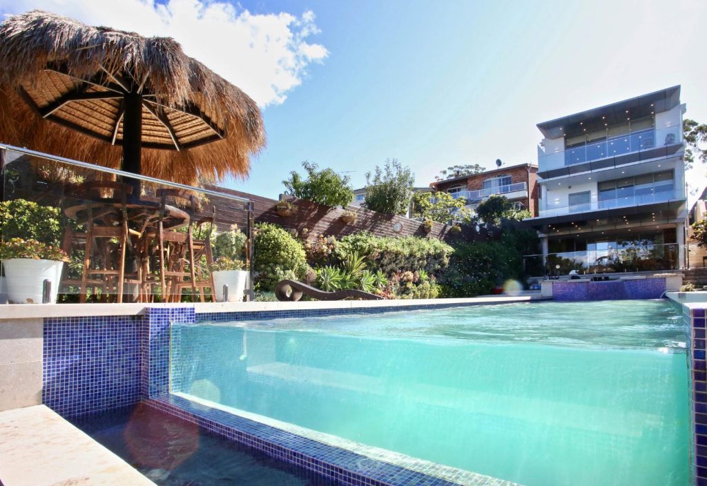 Infinity edge pool with raised glass wall and tropical landscaping in a resort style backyard