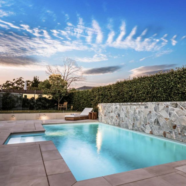 Modern inground concrete pool by Blue Haven Pools with stone wall finish and sunset sky.